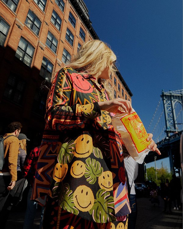 Mulher jovem e branca, de cabelo louro longo, posando para foto nas ruas de Nova York, nos Estados Unidos. Ela usa um vestido longo com estampa de emojis sorrindo coloridos.