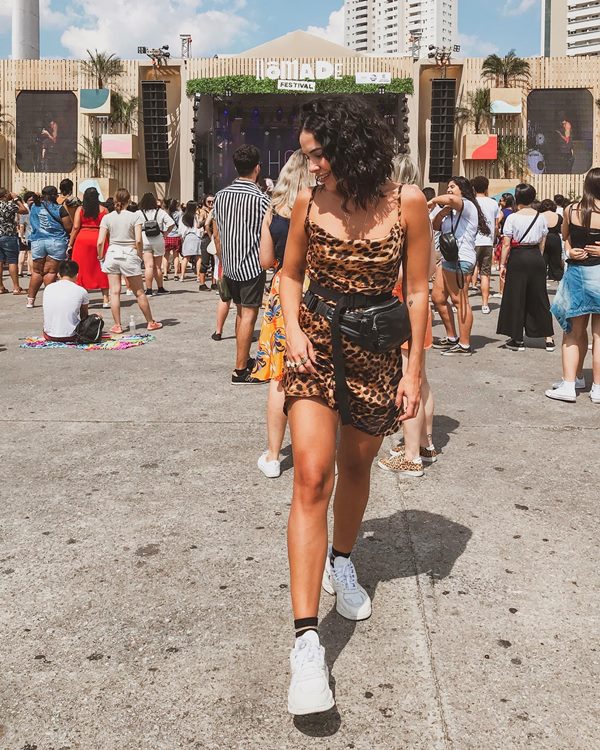 Mulher morena e jovem, de cabelo cacheado curto, posando para foto em um festival. Ela está em frente ao palco e usa um vestido de oncinha curto, uma pochete preta e um tênis branco