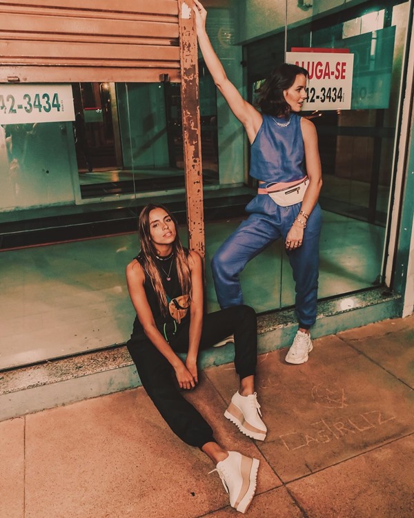 Mulheres brancas e jovem posando para foto em uma área comercial vazia. A primeira possui o cabelo liso e longo e usa um look todo preto com pochete marrom. A segunda possui o cabelo liso curto e usa um conjunto roxo com pochete branca