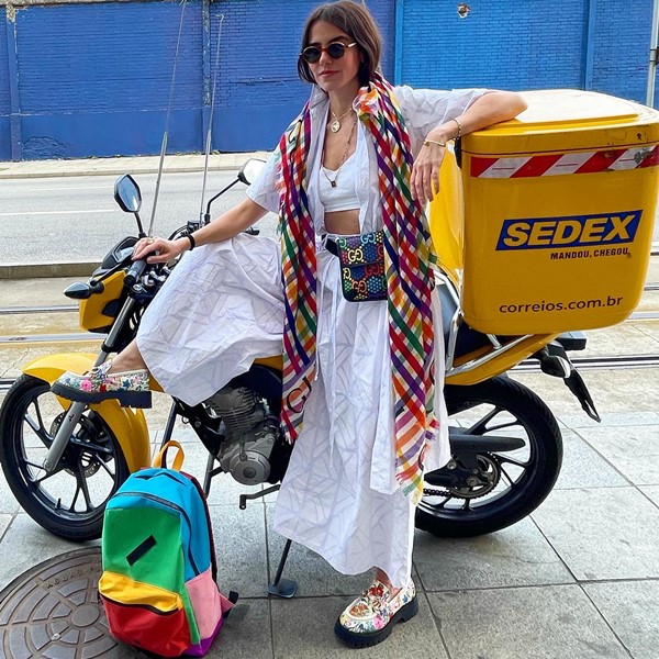 Mulher branca e jovem posando para foto encostada em uma moto amarela do Sedex, serviço de entregas do correio. Ela usa um top, um kimono e uma calça, ambos brancos e fluídos, um cachecom quadriculado colorido e uma pochete preta com desenhos coloridos da marca Gucci.