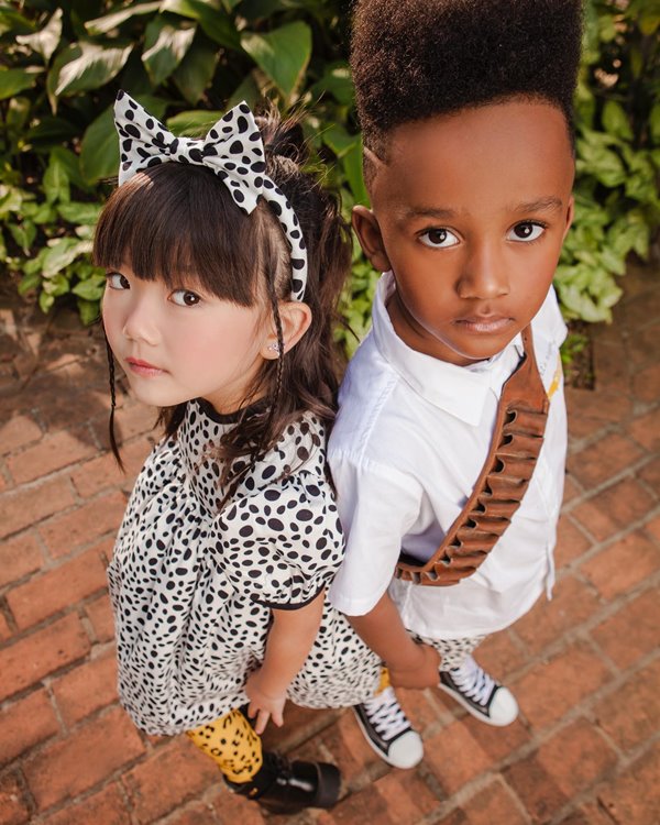 Foto de campanha da marca Sato Kid, fundada pela apresentadora Sabrina Sato. Na foto é possível ver duas crianças usando roupas da marca. A menina é asiática, com o cabelo liso, e usa um vestido com estampa preta e branca; o menino é negro com cabelo curto cacheado e usa uma camisa branca e tênis preto all star.