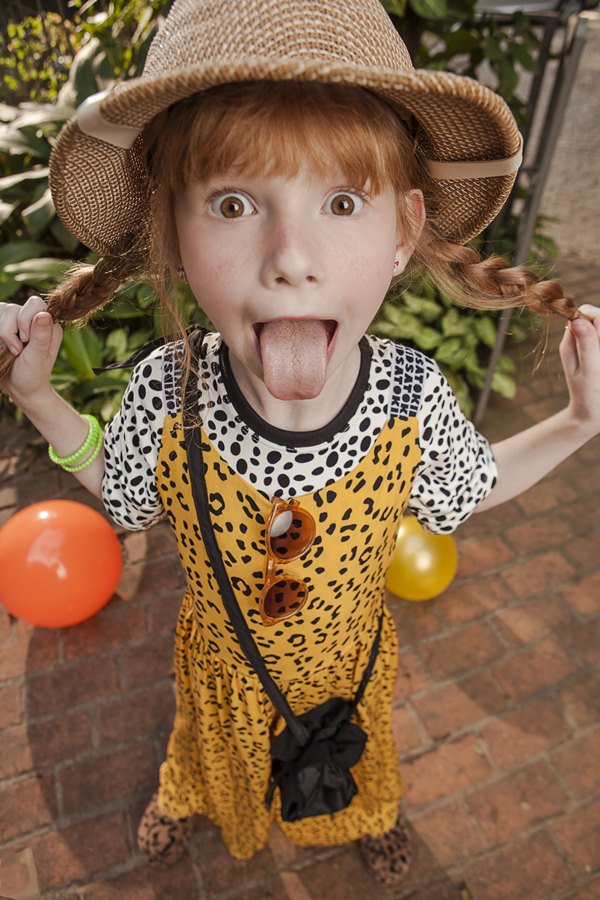 Foto de campanha da marca Sato Kid, fundada pela apresentadora Sabrina Sato. Na foto é uma menina branca e ruiva, criança, usa uma camiseta de zebra e, por cima, um vestido com estampa de onça. Usa ainda uma bolsa preta transpassada e um chapéu estilo safari