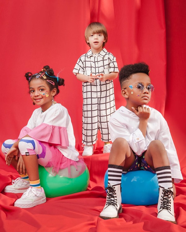 Três crianças posando para foto em um fundo vermelho. A imagem é da campanha publicitária da marca Sato Kid. A primeira é uma menina negra com cabelo cacheado, que veste camiseta e legging, o segundo é um menino branco e louro de mamacão preto e branco, e o terceiro é um menino negro de cabelo cacheado que usa bota coturno e camisa de botão branca