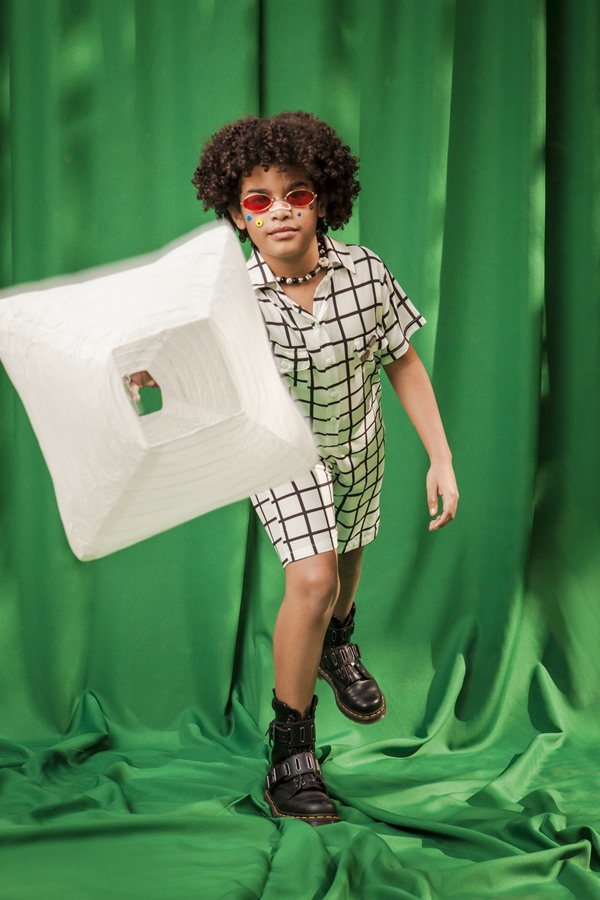 Menino negro de cabelo cacheado, que deve ter por volta de cinco anos, posando para foto em um fundo verde. Ele usa um conjunto de shorts e camisa de botão, ambos brancos com listras pretas, e um óculos de sol com lente vermelha