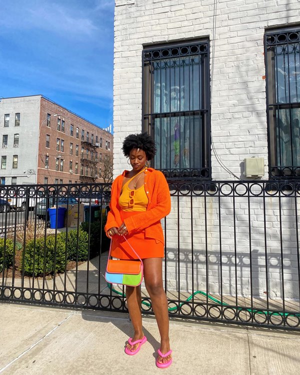 Mulher jovem e negra, com cabelo cacheado curto, posando para foto nas ruas de Nova York, nos Estados Unidos. Ela usa um top curto, uma saia jeans e uma jaqueta, todas as pelas em laranja, uma bolsa retangular de couro colorida e uma rasteira acolchoada rosa da marca Melissa.