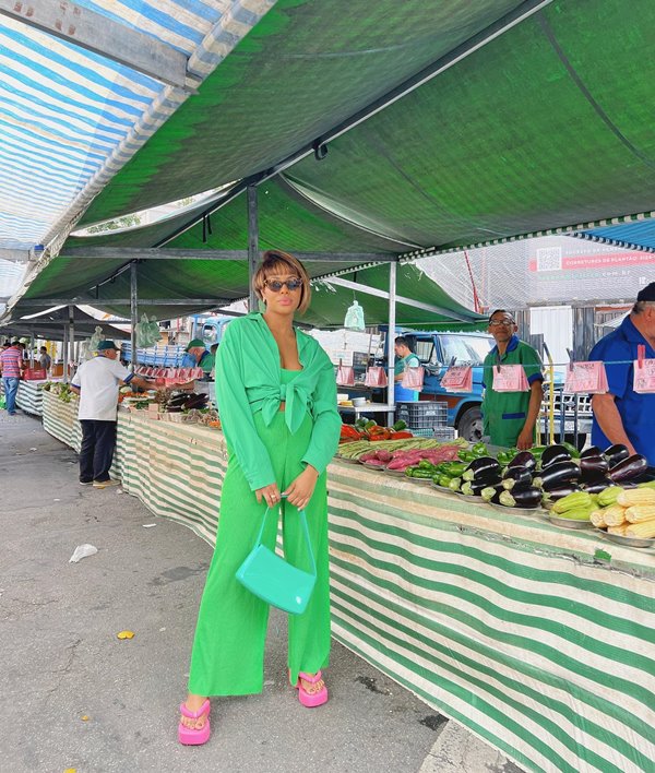 Mulher jovem e negra, com cabelo curto liso, posando para foto ao lado de uma barraca de feira. Ela usa um camisa de botão amarrada em um nó frontal, calça larga e uma bolsa, e todas as peças são verdes, menos o sapato, que é um chinelo plataforma na cor rosa.