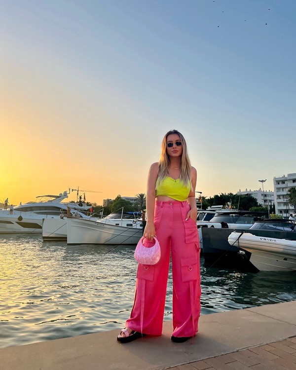 Mulher branca e jovem, de cabelos lisos e loiros, posando para foto no deck de madeira de uma praia. Ela usa um top de seda amarelo, uma calça folgada rosa, e sandália e bolsa na mesma cor.