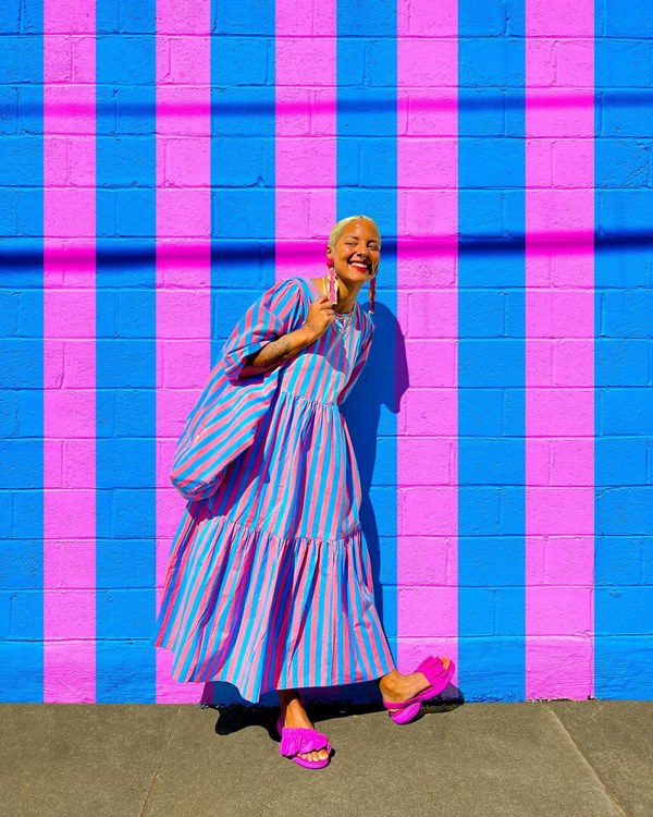 Mulher jovem e negra, de cabelos raspados e loiros, posando para foto em um muro pintado em listras rosas e azuis. Ela usa um vestido longo e com mangas bufantes listrado, nas mesmas cores da parede, uma bolsa saco estilo ecobag na mesma estampa do vestido e uma sandália rasteira rosa.