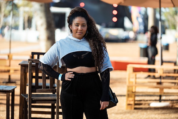 Mulher negra e jovem, de cabelo cacheado longo, posando para foto no Festival CoMA, em Brasília. Ela usa um top preto de renda e, por cima, um top cropped cinza de moletom, calça preta de veludo e uma bolsa também preta.