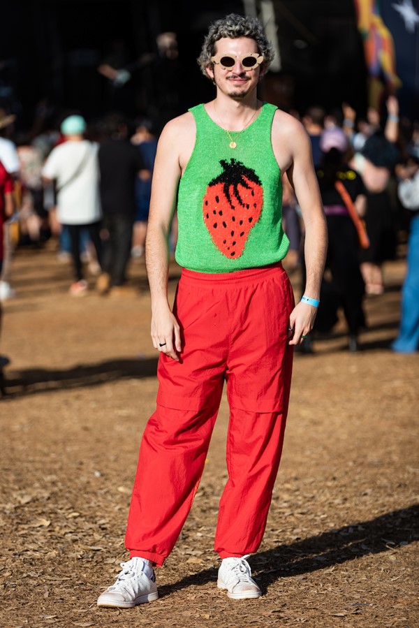 Homem branco e jovem, de cabelo liso descolorido, posando para foto no Festival CoMA, em Brasília. Ele usa um top de tricô verde com um morango grande estampado no centro, uma calça vermelha e um óculos escuro com lentes em formato de flor