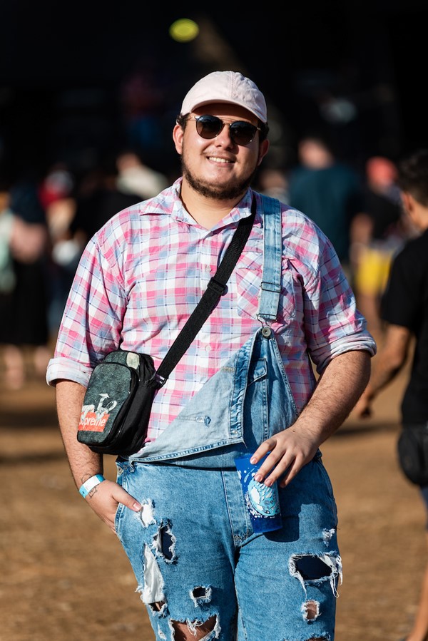 Homem branco e jovem, de cabelo curto preto, posando para foto no Festival CoMA, em Brasília. Ele usa óculos escuros, boné rosa, uma camisa de botão rosa e uma jardineira jeans.