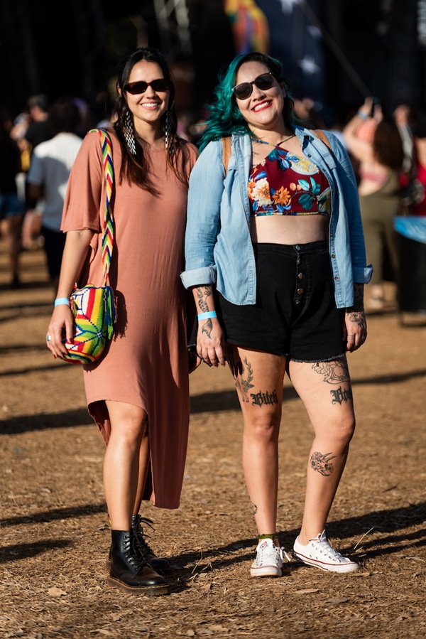 Mulheres brancsa e jovens posando para foto no Festival CoMA, em Brasília. A primeira tem o cabelo liso louro e usa um vestido rosa, uma bolsa colorida e botas coturno. A segunda tem o cabelo azul ondulado, usa um top colorido, um shorts preto e uma camisa jeans por cima