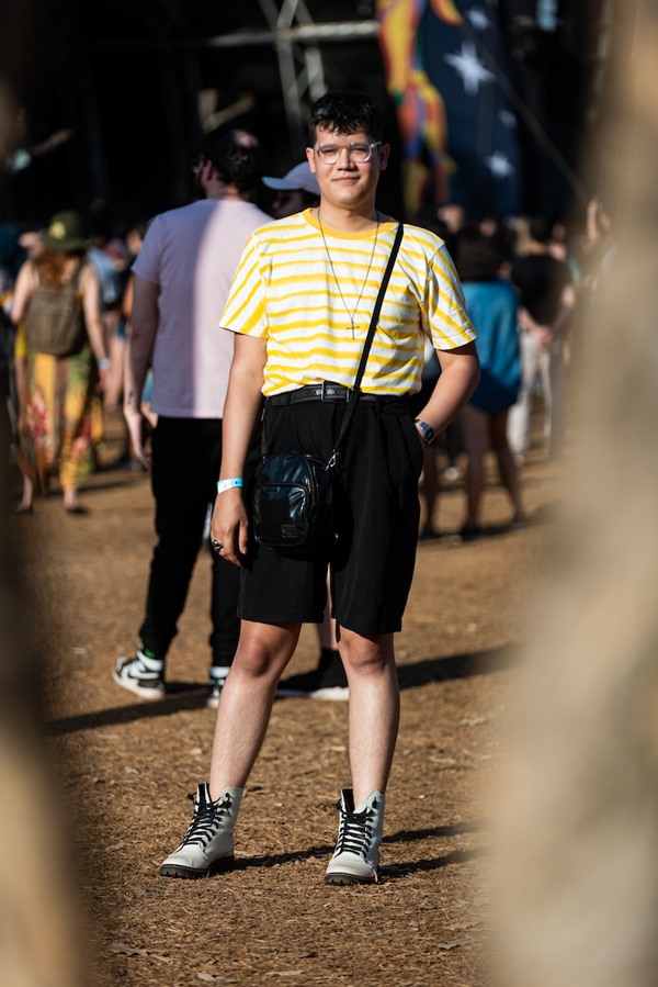 Homem branco e jovem, de cabelo liso preto, posando para foto no Festival CoMA, em Brasília. Ele usa óculos de grau, uma camiseta amarela e branca listrada, bermuda preta, uma bolsa preta transpassada e coturno branco e preto