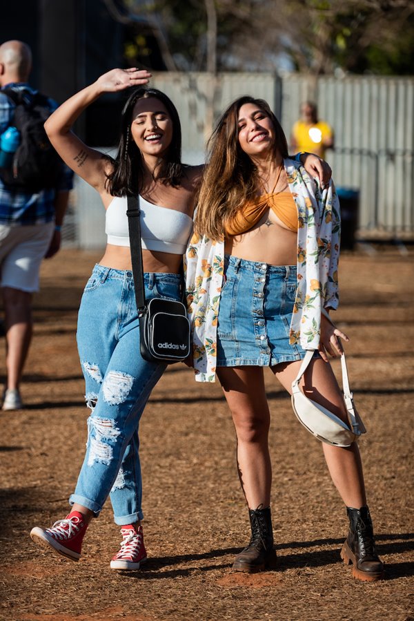 Mulheres brancas e jovens posando para foto no Festival CoMA, em Brasília. A primeira tem o cabelo liso curto e preto e usa um top branco, calça jeans rasgada e tênis all star vermelho. A segunda tem o cabelo longo castanho com mechas loiras e usa um top amarelo, saia jeans e um kimono estampado