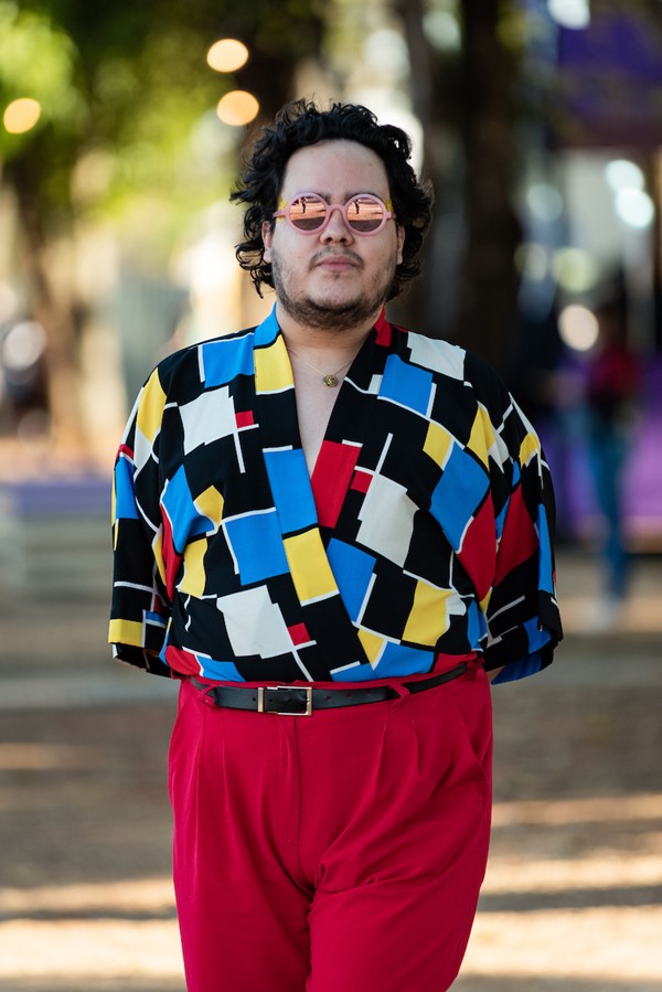 Homem branco e jovem, de cabelo liso preto, posando para foto no Festival CoMA, em Brasília. Ele usa óculos de escuros com lente rosa, uma camisa colorida com quadrados pretos, branco, azul e vermelho e uma calça vermelha