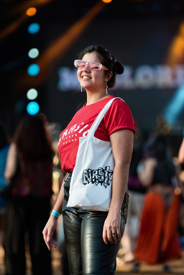 Mulher branca e jovem, de cabelo liso preto, posando para foto no Festival CoMA, em Brasília. Ela usa uma camiseta vermelha escrito BRASIL em branco, uma calça de couro preta, uma sacola estilo ecobag branca e óculos com lente rosa