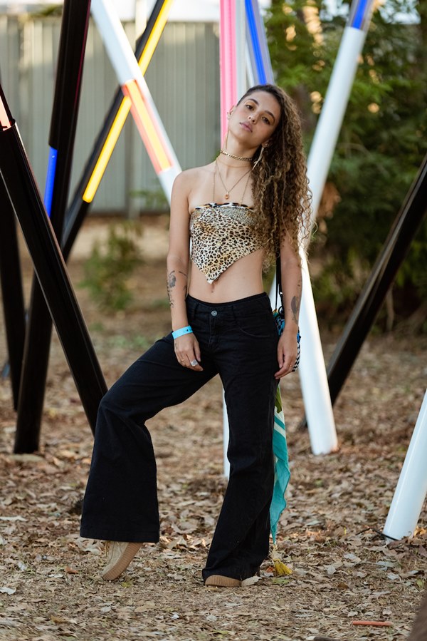 Mulher branca e jovem, de cabelo grande loiro e cacheado, posando para foto no Festival CoMA, em Brasília. Ela usa um lenço de onça amarrado como top, calça jeans e tênis bege