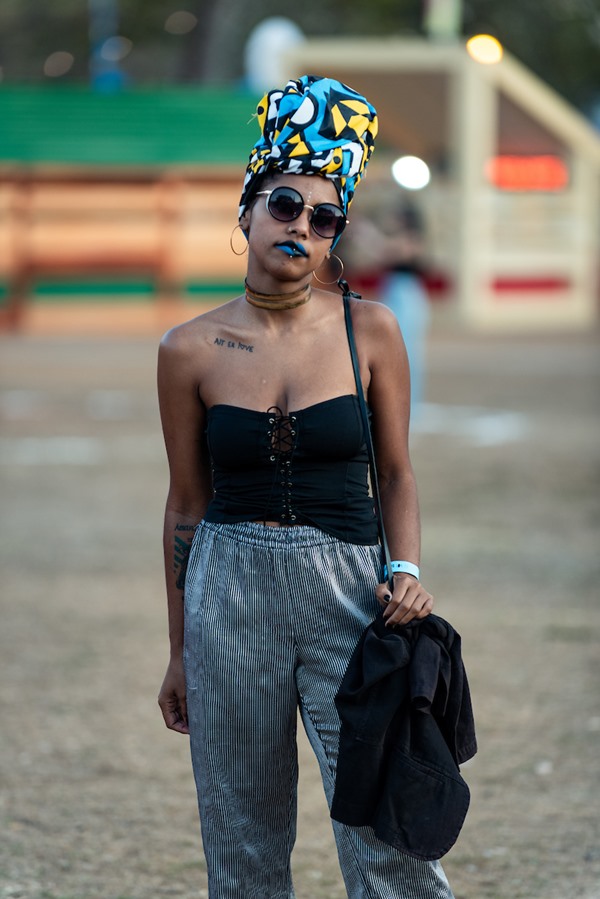 Mulher negra e jovem, de turbante colorido e óculos escuros, posando para foto no Festival CoMA, em Brasília. Ela um top preto, calça cinza e uma bolsa preta no ombro