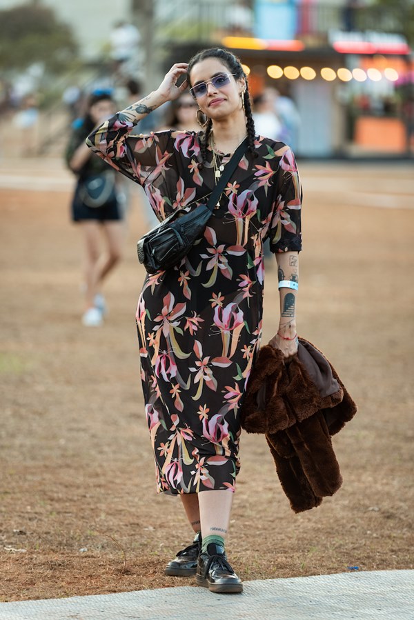 Mulher morena e jovem, de cabelo amarrado em duas tranças, posando para foto no Festival CoMA, em Brasília. Ela usa um vestido longo estampado com tecido transparente, tênis preto, óculos escuros e uma pochete preta transpassada.