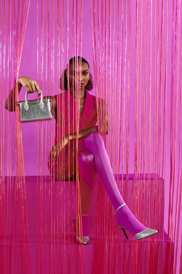 Mulher jovem e negra, com cabelo cacheado amarrado em um rabo de cavalo baixo, posando para foto na campanha da marca Corello. Ela está em um fundo rosa e usa um um vestido rosa, uma meia calça rosa, e uma sandália de salto e uma bolsa totte, ambas prata.