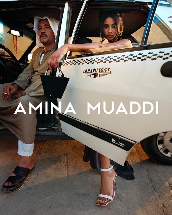 Campanha de divulgação da marca de sapatos Amina Muaddi. Na foto, é possível ver uma mulher sentada no banco de trás de um taxi branco com uma sandália de salto prata e uma bolsa pequena preta de couro. Na frente, no banco do motorista, tem um homem árabe usando roupas típicas 