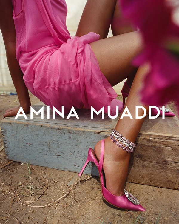 Campanha de divulgação da marca de sapatos Amina Muaddi. Na foto, é possível ver as pernas de uma mulher morena que usa um vestido rosa e uma sandália de salto, também rosa, com pedras estilo diamante