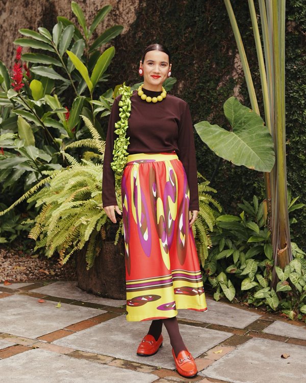 Mulher branca e jovem, de cabelos lisos amarrados, posando para foto em um jardim. Ela usa uma blusa marrom, uma saia colorida, sapatos mocassim de couro vermelho e um colar de contas amarelo