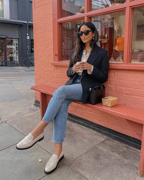 Mulher jovem e negra, de cabelos lisos longo, posando para foto sentada na entrada de um café em Paris. Ela usa óculos escuros, uma camisa de seda branca, um blazer azul, calça jeans e um sapato mocassim de couro branco com preto