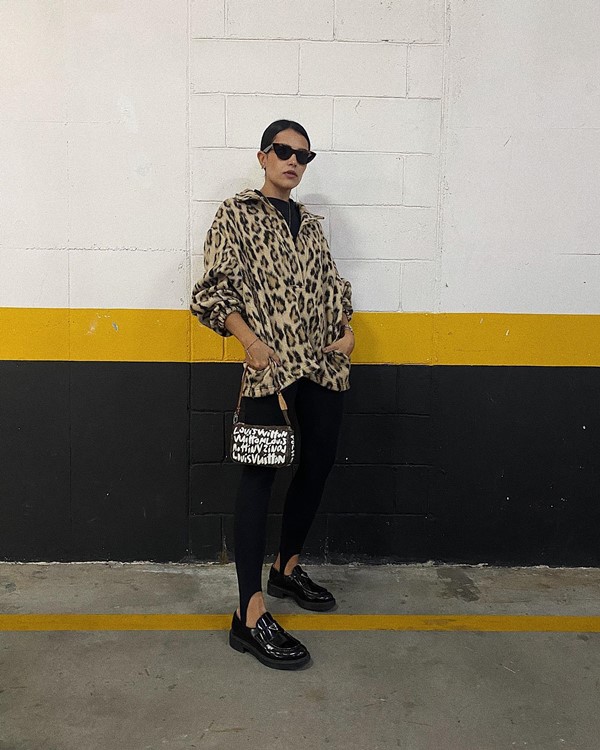 Mulher jovem e branca, de cabelos lisos castanhos amarrados, posando para foto em um estacionamento. Ela usa óculos escuros, uma camiseta preta, um casaco de pelinho com estampa de onça, calça legging preta e um mocassim preto de couro