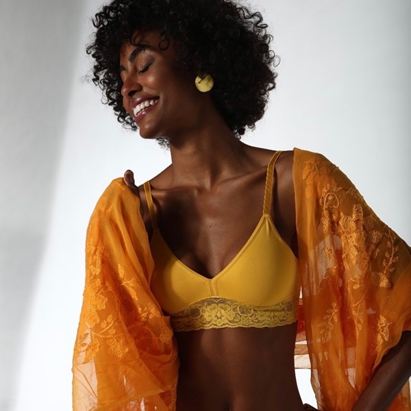 Mulher jovem e negra, com cabelo curto cacheado, posando para foto em um fundo branco e usando um sutiã amarelo e um kimono laranja por cima