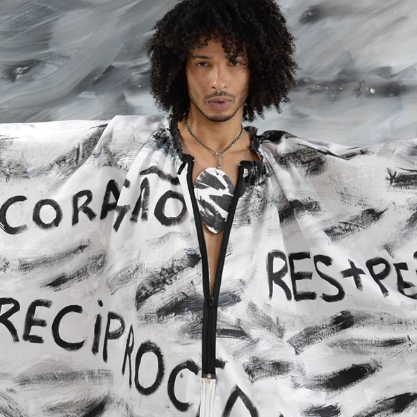 Modelo homem negro e jovem, com o cabelo cacheado médio, posando para foto em um fundo preto e branco. Ele usa um vestido túnida, também preto e branco, com as palavras CORAÇÃO, RESPEITO E RECIPROCIDADE escritas.