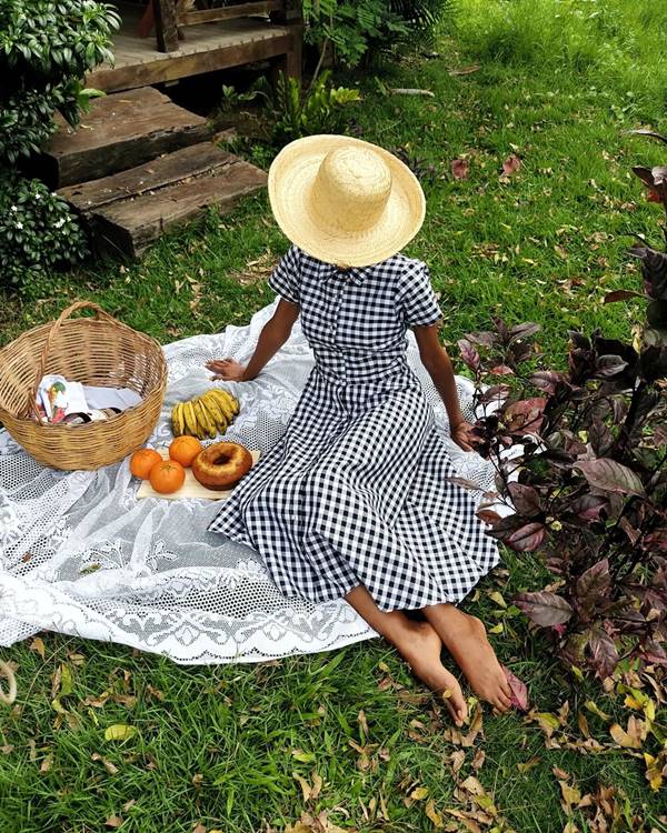 Mulher jovem e negra, sentada em uma toalha de piquenique, usando um vestido quadriculado preto e branco e um chapéu de palha. 