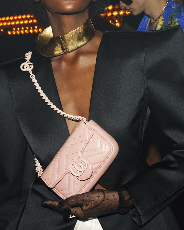 Mulher jovem e negra, que usa um blazer preto decorado e um colar dourado, segurando bolsa rosa clara da marca Gucci