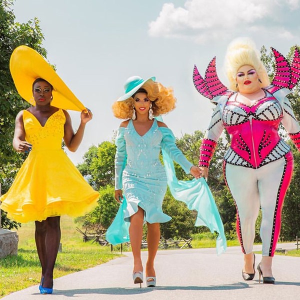 Três drag queens participantes da série We're Here, da HBO. Na foto, as três andam por uma rua com roupas extravagantes. A primeira é negra e usa um vestido e um chápeu amarelo, a segunda também é negra e usa um vestido e chápeu azul claro e a terceira é branca, com peruca loura, e usa um macacão colado branco e rosa