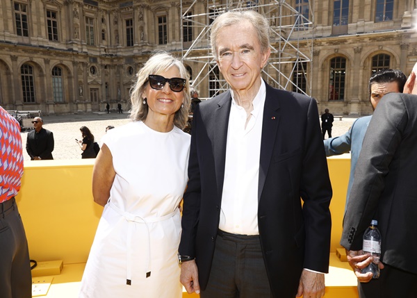 O empresário francês Bernard Arnault, um homem idoso e branco, de cabelo liso, branco e curto, posando para foto no desfile da Louis Vuitton, marca da qual é dono. Ele usa camisa branca de botão, um blazer escuro e gravata. Ao seu lado está uma mulher loura, de pele bronzeada e meia idade. É sua esposa, Hélène Mercier, que veste um vestido branco e óculos escuros na cor preta.