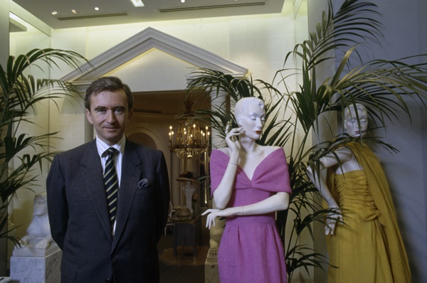 O empresário francês Bernard Arnault, um homem idoso e branco, de cabelo liso, branco e curto, posando para foto em frente a manequins com roupas da marca Dior. Ele usa camisa branca de botão, um blazer escuro e gravata. A foto é de 1988.