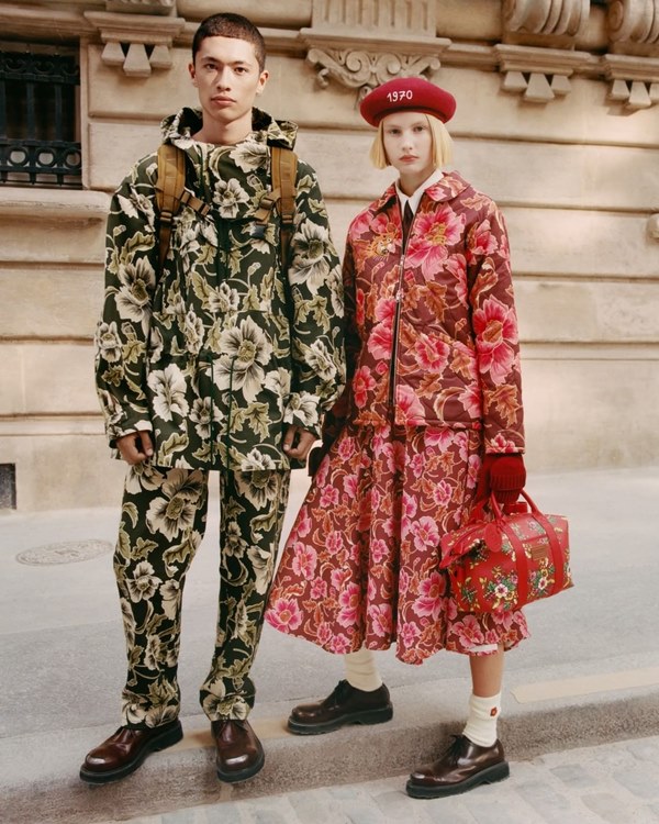 Foto de campanha da marca de luxo Kenzo com dois modelos brancos nas ruas de Paris. Ele, de cabelo castanho raspado, usa um conjunto de calça e casaco com capuz na mesma estampa floral verde. A mulher, de cabelo curto, liso e loiro, usa uma boina vermelha e um conjunto de casaco com saia, ambos em uma estampa rosa floral.