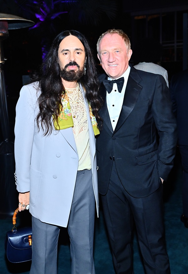 O estilista italiano Alessandro Michele, um homem jovem, branco, de barba e cabelos longos castanhos, posando ao lado do empresário François-Henri Pinault, um homem idoso, branco e de cabelos também brancos