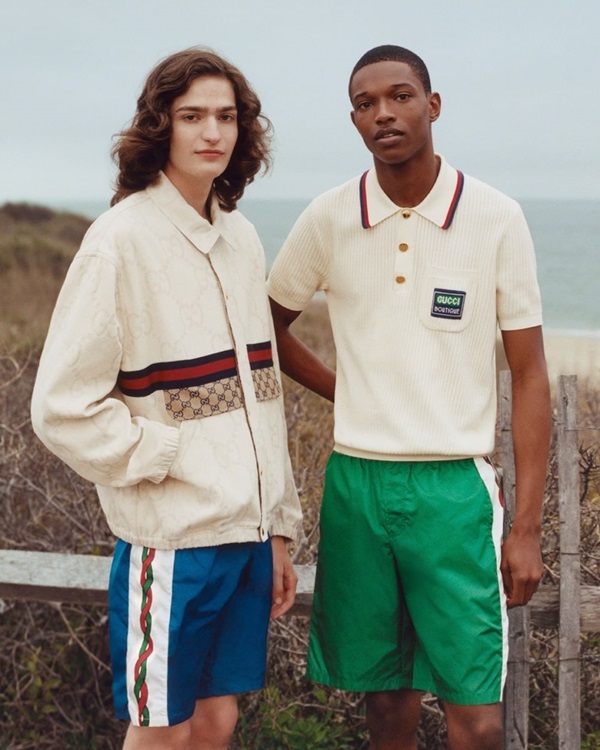 Campanha da linha de roupas masculinas da marca italiana Gucci. Os dois modelos, um homem branco de cabelo grande, e um negro de cabelo raspado, posam para foto em uma praia. O primeiro usa um casaco de moletom bege e um shorts azul; e o segundo usa uma camisa polo, também bege, e um shorts verde