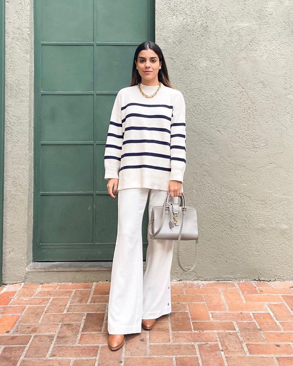 Mulher branca e jovem, de cabelos lisos castanhos, posando para foto na área externa de casa. Ela usa uma calça branca de boca de sino, um casaco branco com listras preta, um salto alto bege e uma bolsa cinza de alças longas