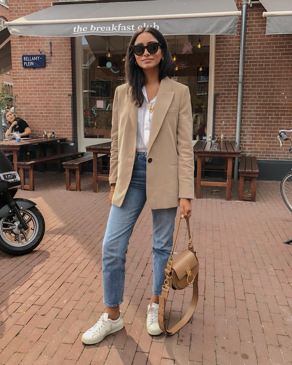 Mulher jovem e morena, de cabelos lisos longos, posando para foto em frente a um café em Paris. Ela usa umacamisa de botão branca, calça jeans azul, um blazer bege por cima e, nos pés, um tênis de couro branco. Em uma das mãos ela segura uma bolsa de couro na cor caramelo da marca Dior.