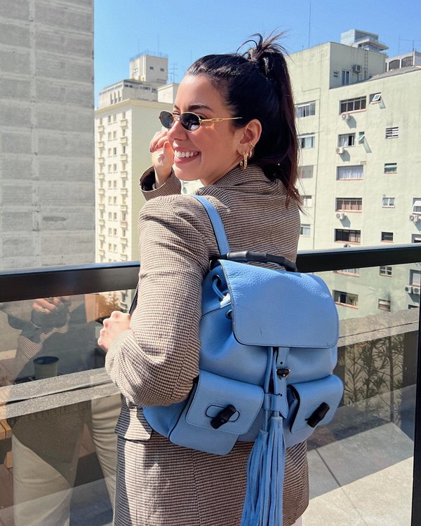 Mulher branca e jovem, de cabelos lisos castanhos amarrados em um rabo de cavalo, posando para foto na varanda de um prédio em São Paulo. Ela usa um blazer quadriculado marrom, óculos de sol e uma mochila de couro azul da marca Gucci