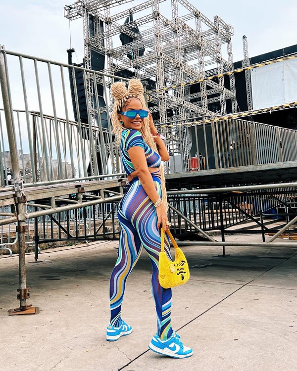 Mulher negra e jovem, de cabelos loiros trançados longos, posando para foto em um festival de música. Ela usa um conjunto de blusa e legging, com estampa azul, um tênis azul claro da Nike e uma bolsa amarela de textura acolchoada da Prada