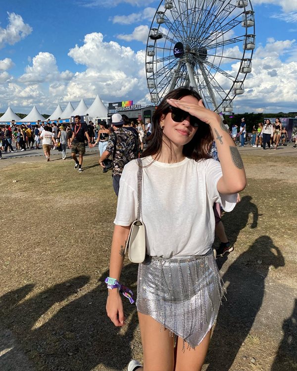 Mulher branca e jovem, com cabelo longo ondulado e castanho, posando para foto no festival Lollapalooza. Ela veste uma camiseta branca, uma saia brilhante prateada e uma bolsa de couro branca, além de óculos escuros