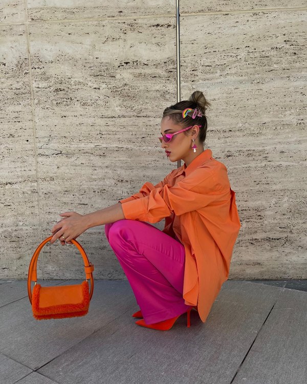 Mulher branca e jovem, de cabelo liso loiro, posando para foto agachada na área externa da própria casa. Ela está com uma camisa laranja de botão, uma calça rosa, sandália de salto e bolsa, ambas laranjas