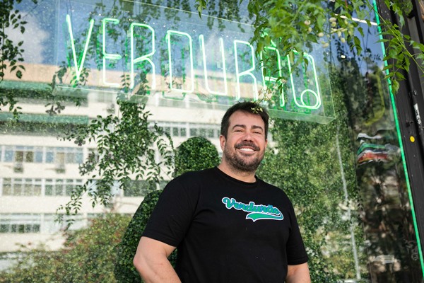 Ensaio fotográfico feito na loja da marca Verdurão Camisetas, na W3 Sul, em Brasília. Na foto é possível a fachada do estabelecimento e Wesley Santos, dono e diretor da marca. Ele é um homem branco jovem, de cabelo castanho, que usa uma calça jeans e uma camiseta preta com a logomarca da Verdurão