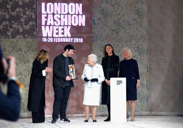 A Rainha Elizabeth II durante a Semana de Moda de Londres em 2018. A monarca usa um casaco azul claro de tweed, luvas e sapatos de salto baixo, ambos em couro preto. Ela entrega um prêmio ao estilista Richard Quinn, um homem jovem, branco, que usa camiseta, casaco e calça, tudo na cor preta.