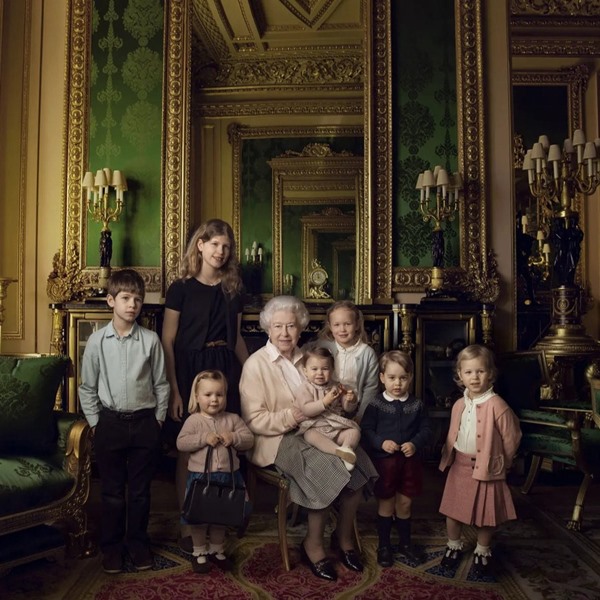 A Rainha Elizabeth II, em 2017, durante uma sessão de fotos para a revista Vogue. Ela está em uma sala do castelo rodeada pelos seus netos e bisnetos. Usa uma camisa branca de botão, um cardigã rosa claro, uma saia cinza e sapatos estilo mocassim preto e de couro.