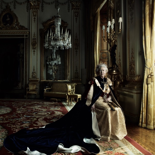 A Rainha Elizabeth II, em 2017, durante uma sessão de fotos para a revista Vogue. Ela está sentada em uma cadeira na varanda da sala do castelo e usa uma capa clássica de reis e a coroa britânica.