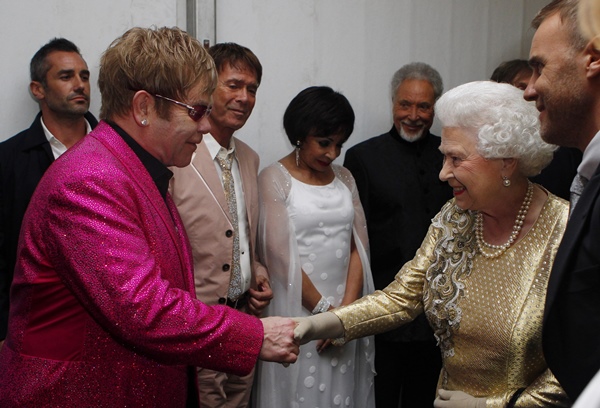 O cantor Elton John, um homem branco e idoso de cabelo loiro liso, apertando as mãos da rainha Elizabeth. O encontro aconteceu em 2012. Ele usa uma camisa de botão preta e um blazer rosa choque com brilho. A monarca usa um vestido dourado com rendas e um colar de pérolas.